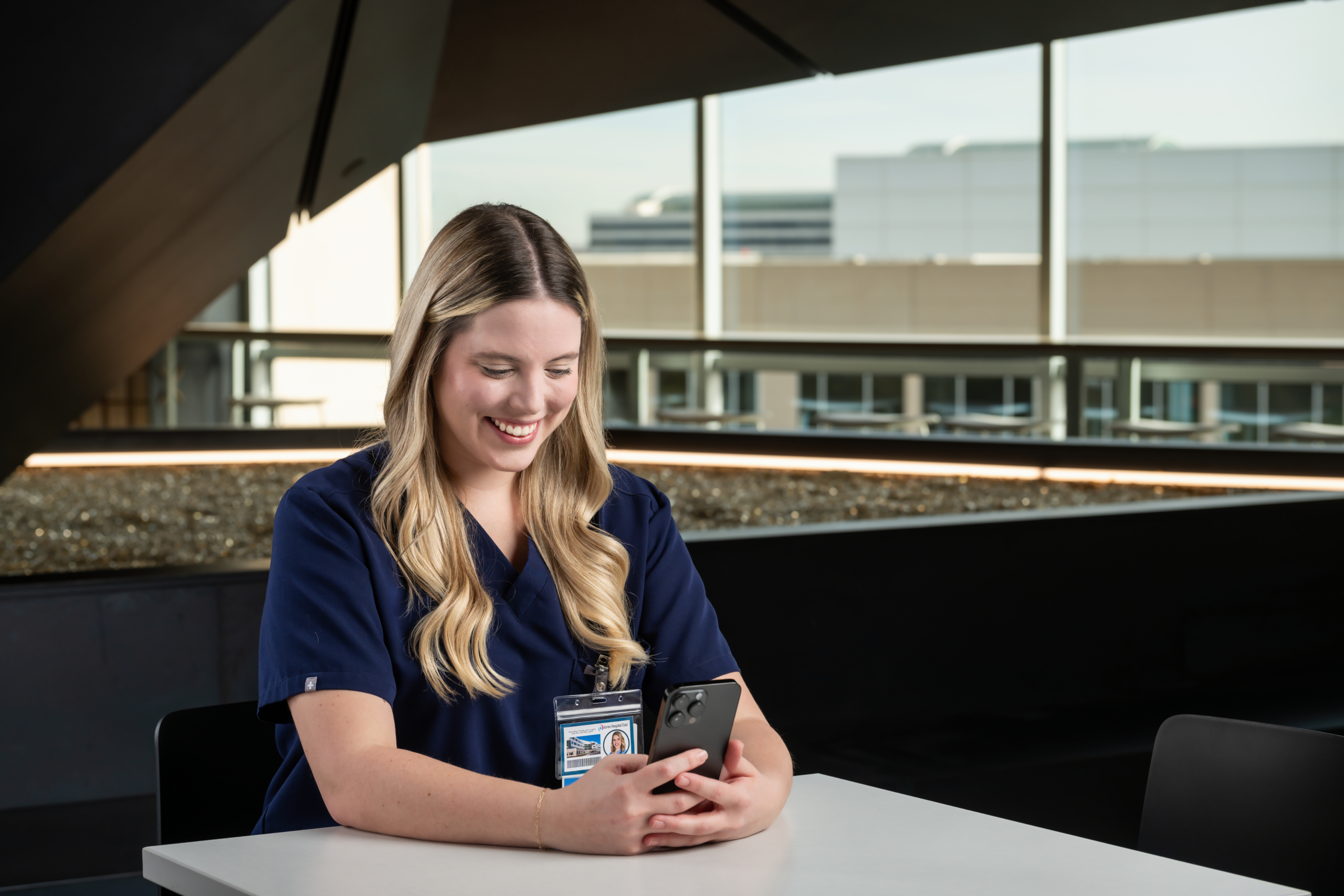 Nurse seated at table looking at phone and smiling.