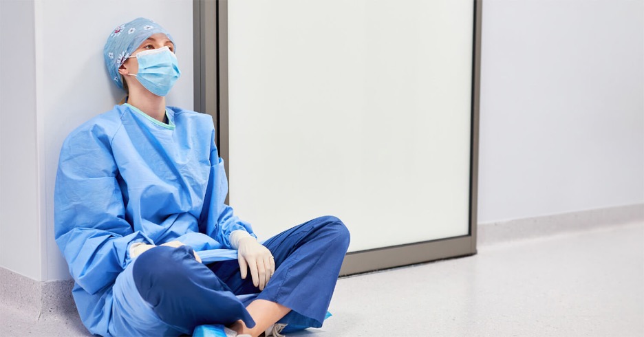 Exhausted nurse with face mask and hair bonnet resting at the end of a hall by sitting on the floor