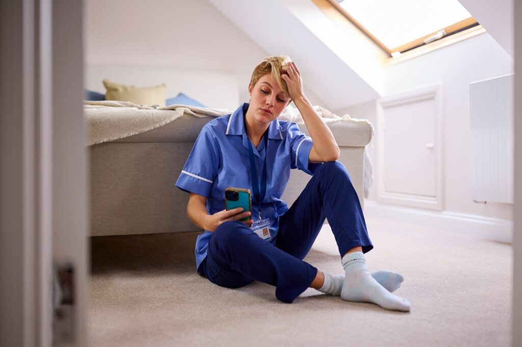 Thoughtful Woman Wearing Nurses Or Doctors Uniform Sitting On Floor At Home Looking At Mobile Phone