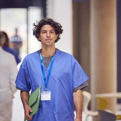 Male Doctor Wearing Scrubs Carrying Patient Notes Walking In Busy Hospital #image_title